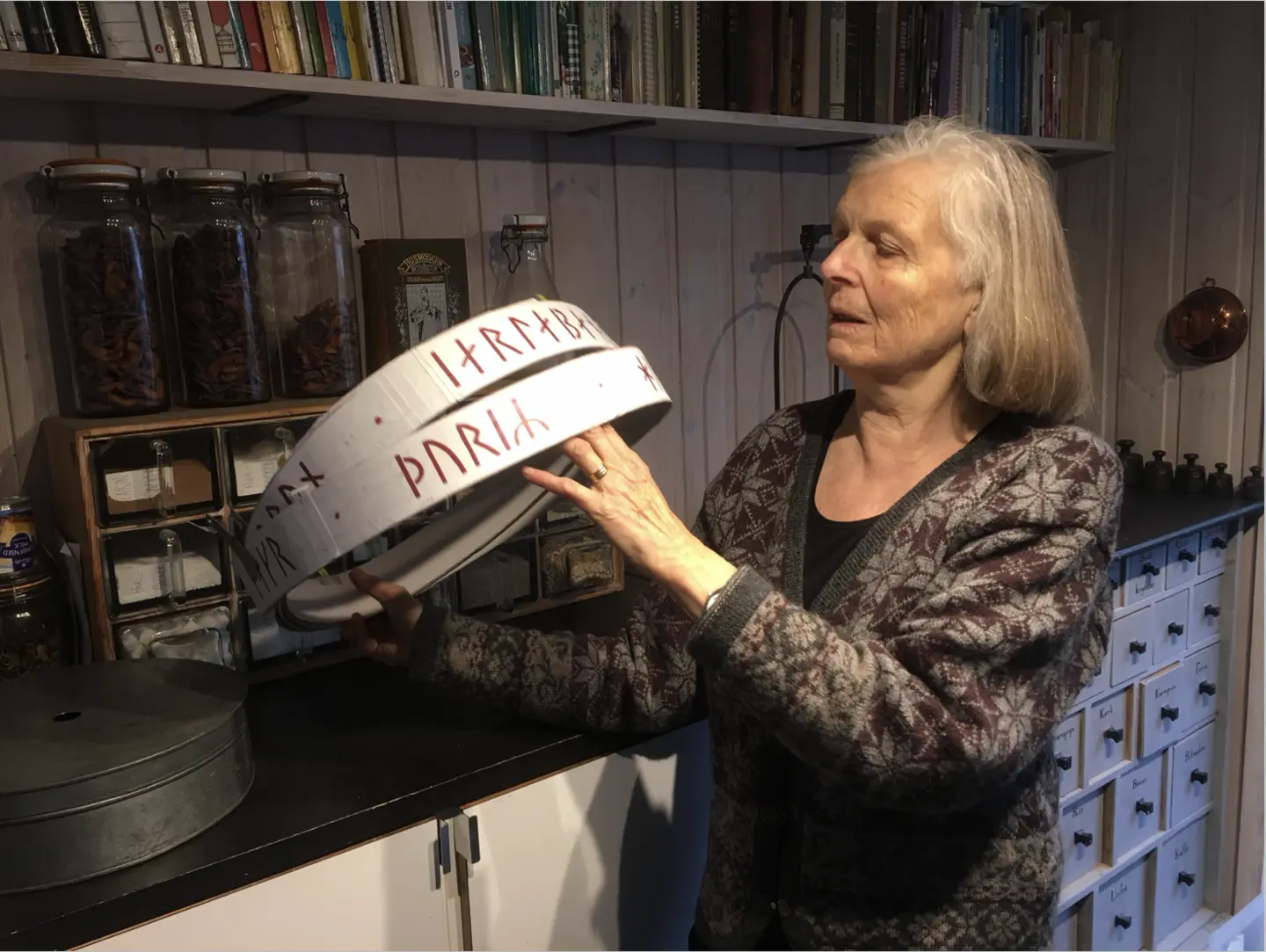 Karin Walde håller en vit modellring med röda runtecken framför hyllor med burkar och böcker.